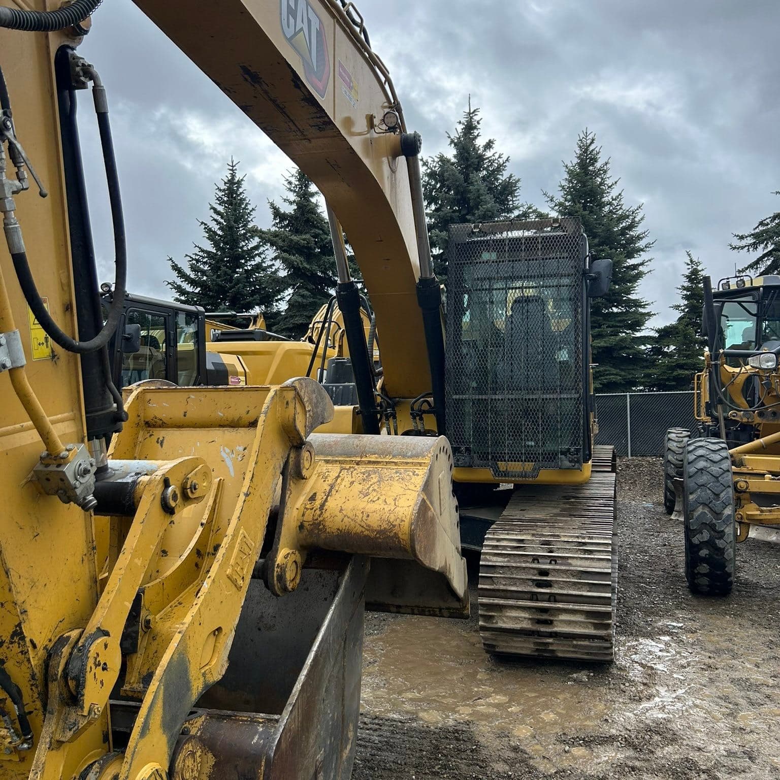 Yellow Caterpillar excavator on muddy ground with evergreen trees under a cloudy sky.